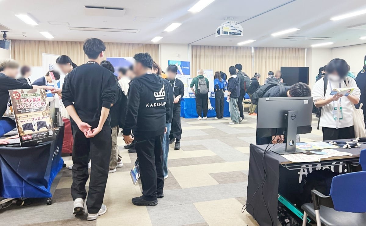 スポンサーブースが並ぶ教室の様子、いくつかの企業のブースを聞きに集まる参加者の背中の写真