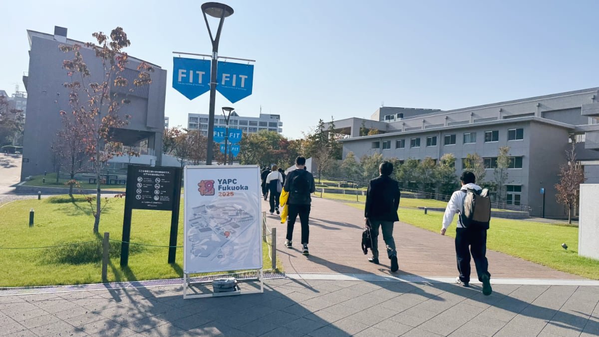 福岡工業大学のキャンパス入り口、YAPC::Fukuoka 2025の看板があり、数名の参加者が会場に向かう背中の写真